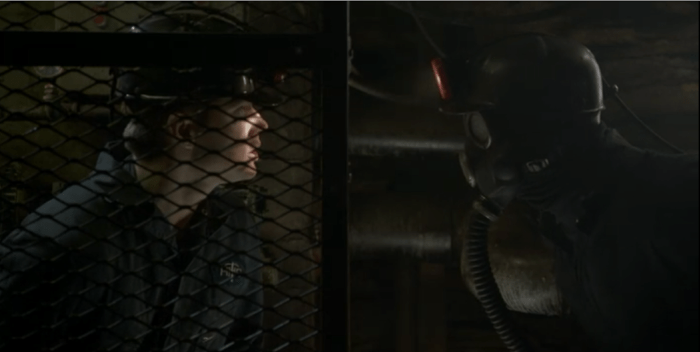 a miner stares at another miner whose face is obscured by a helmet that covers their face