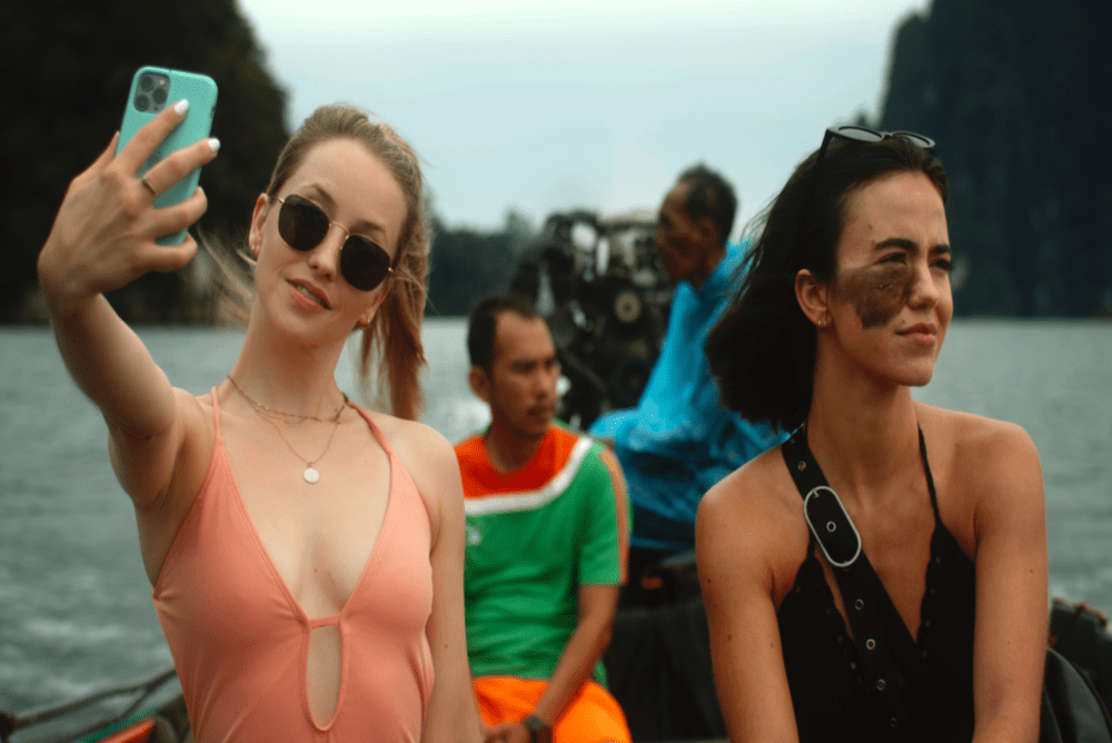 One woman takes a selfie while riding on a boat while another woman looks at the scenery