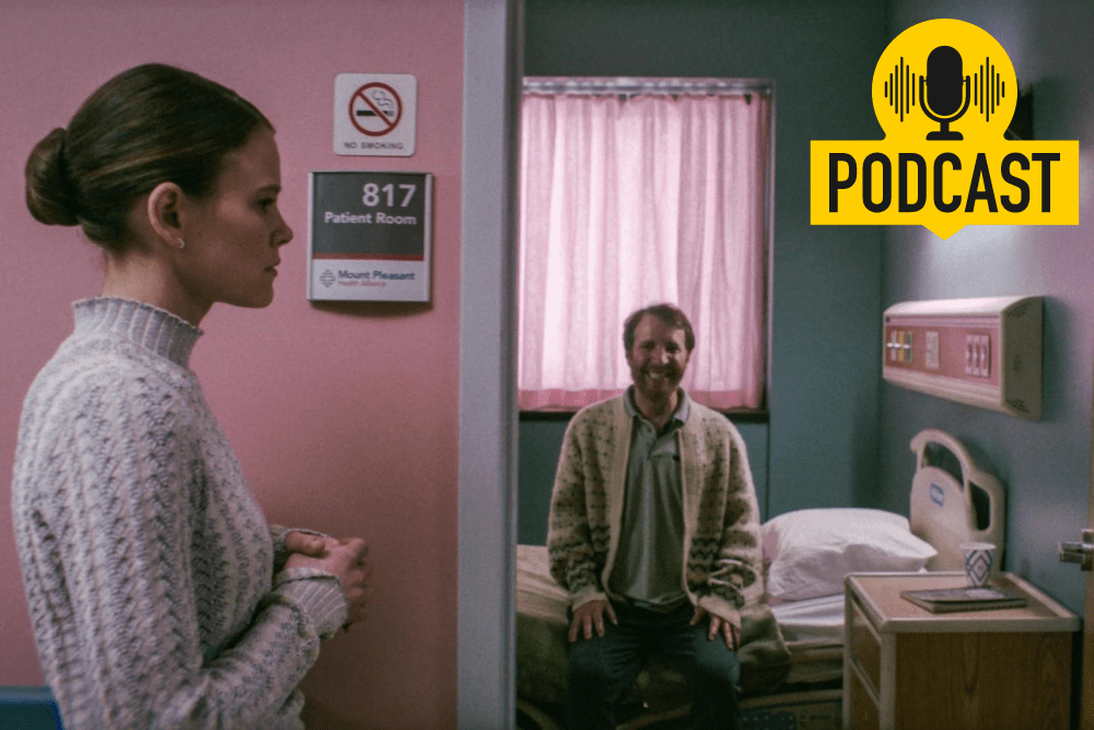A woman stands outside of a door pausing before entering a room. Inside, a man is sitting on a bed and he is smiling.